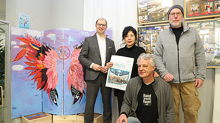 Eine Frau und drei Männer, von denen einer kniet, haben sich vor einem Kunstwerk und mehreren Bilderrahmen mit Fotos in einem großen Raum aufgestellt. Die Frau hält zudem ein Plakat in Händen.