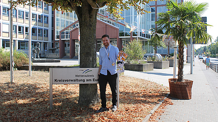 Ein junger Mann steht vor einem Hinweisschild mit der Aufschrift "Wwetteraukreis - Kreisverwaltung Europaplatz"