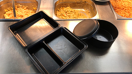 Zwei Mehrwegbehälter für jeweils eine Portion Essen. Im Hintergrund drei Metallbehälter für die Essensausgabe