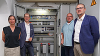 Vier Menschen stehen in einem Keller vor einem Metallschrank, in dem sich viel Technik befindet.