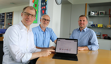 Drei Männer sitzen an einem runden Tisch. Vor einem steht ein aufgeklappter Laptop. Der Mann deutet auf den Bildschirm.