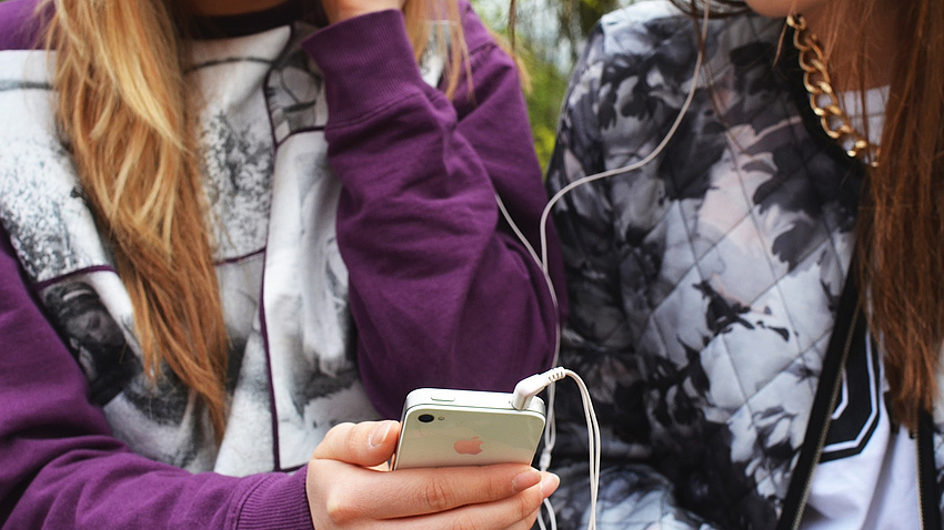 Zwei Jugendliche stehen dicht beieinander und hören gemeinsam über einen Kopfhörer Musik, abgespielt von einem Smartphone.