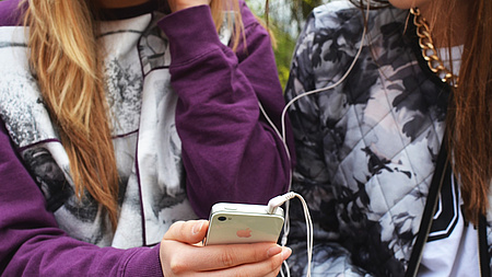 Zwei Jugendliche stehen dicht beieinander und hören gemeinsam über einen Kopfhörer Musik, abgespielt von einem Smartphone.