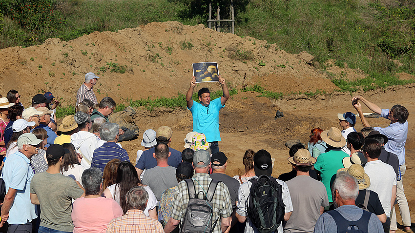 Eine größere Personengruppe steht im Halbkreis um einen Mann, der ein großformatiges Foto hochhält.