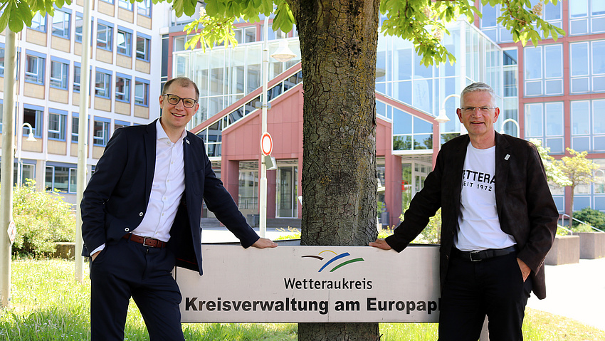 Zwei Männer stehen neben einem Schild mit der Aufschrift "Wetteraukreis. Kreisverwaltung am Europaplatz"