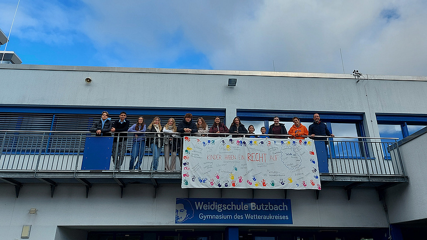 Mehrere Personen stehen auf dem Balkon eines Gebäudes. Vom Geländer hängt ein großes Plakat herab, auf dem Statements zu Rechten von Kindern stehen.
