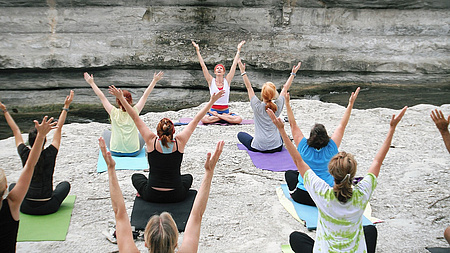 Eine sitzende Gruppe Frauen auf einem Fels am Fluss mach Yoga und streckt die Arme Richtung Himmel