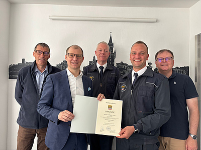 Fünf Männer, zwei von ihnen in Feuerwehruniform, stehen vor einer Wand. Einer der Männer überreicht einem der Uniformierten eine Urkunde.