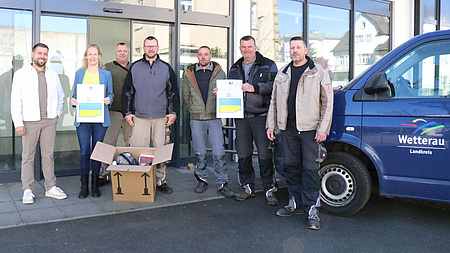 Sieben Personen, sechs Männer und eine Frau, stehen vor einer Glasfassade. Die Frau und einer der Männer halten je ein Plakat mit einer ukrainischen Fahne hoch. Vor ihnen steht ein Karton mit gespendeten Gerätschaften.