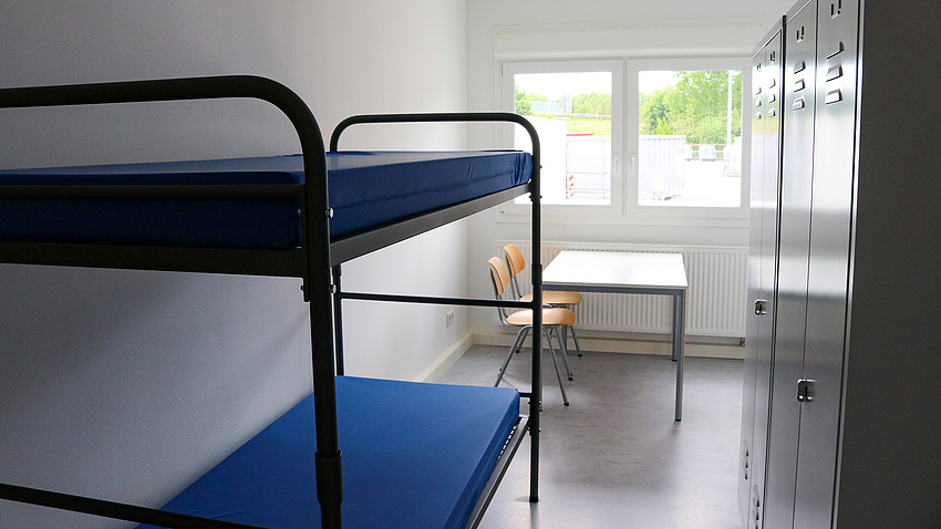 Ein Zimmer mit einem Stockbett und vorne am Fenster einem Tisch, an dem zwei Stühle stehen. Rechts zwei Metallschränke.