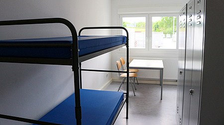 Ein Zimmer mit einem Stockbett und vorne am Fenster einem Tisch, an dem zwei Stühle stehen. Rechts zwei Metallschränke.
