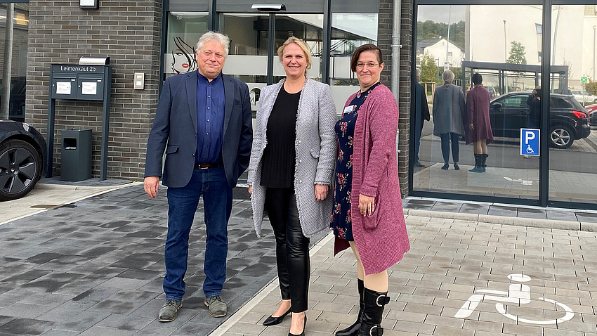 Zwei Frauen und ein Mann stehen vor einem Gebäude. Daran hängt ein Schild mit der Aufschrift "Alloheim".