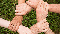 Das Foto zeigt sechs Hände, deren Besitzer jeweils das Handgelenk einer Hand ihres Nachbarn/ihrer Nachbarin umfassen, so dass ein Kreis entsteht.