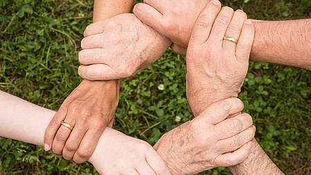 Das Foto zeigt sechs Hände, deren Besitzer jeweils das Handgelenk einer Hand ihres Nachbarn/ihrer Nachbarin umfassen, so dass ein Kreis entsteht.