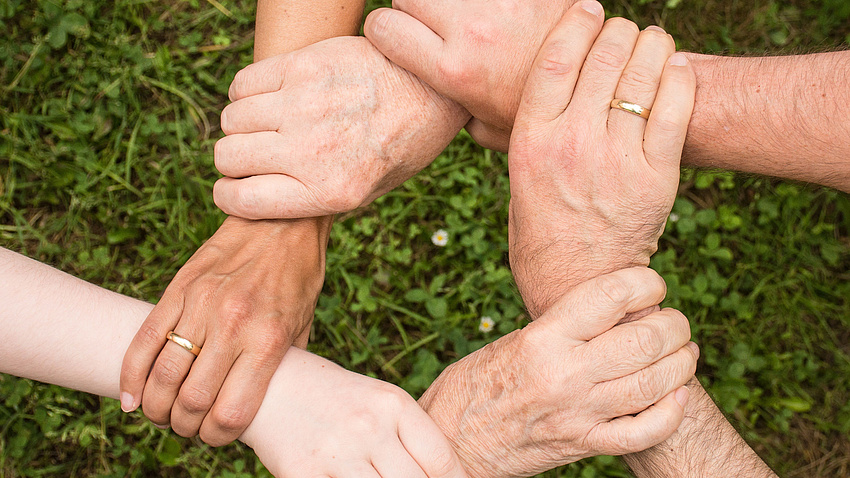 Das Foto zeigt sechs Hände, deren Besitzer jeweils das Handgelenk einer Hand ihres Nachbarn/ihrer Nachbarin umfassen, so dass ein Kreis entsteht.