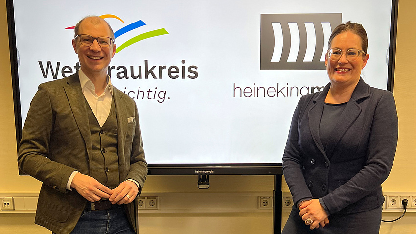 Der Landrat und eine Mitarbeiterin stehen vor einer interaktiven Schultafel und blicken in die Kamera.