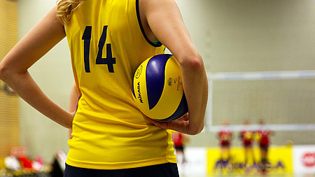 Zu sehen ist eine Person in einem gelben Trikot, die sich einen blau-gelben Volleyball unter den Arm geklemmt hat.