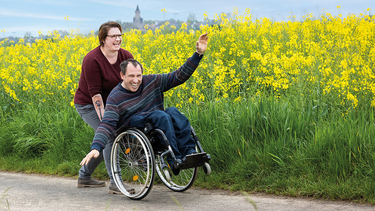 Auf einem Feldweg mit gelben Rapsblüten im Hintergrund sieht man einen Mann im Rollstuhl. Hinter ihm schiebt eine Frau seinen Rollstuhl und hebt die vorderen Rädern auf. Der Mann hat seine Armen ausgestreckt, als ob er fliegt. Beide Menschen strahlen. 