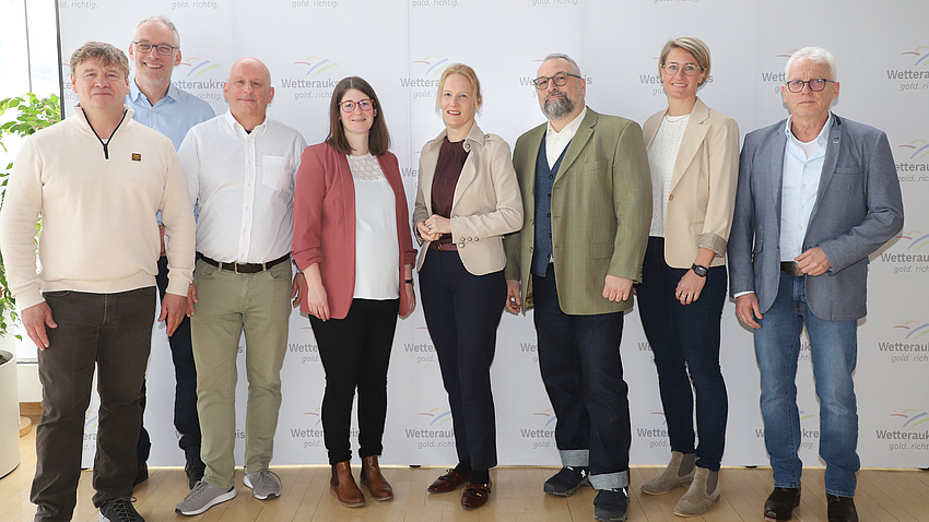 Drei Frauen und fünf Männer stehen in einem größeren Saal vor einer Fotowand.