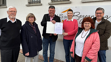 Sechs Leute vor einem Gebäude, an dem ein Schild "Haus der Begegnung" hängt.