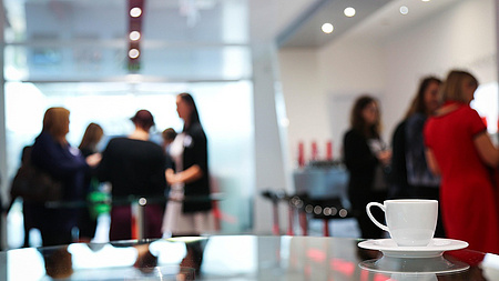Büroraum mit einer Tasse im Vordergrund, im Hintergrund viele Frauen