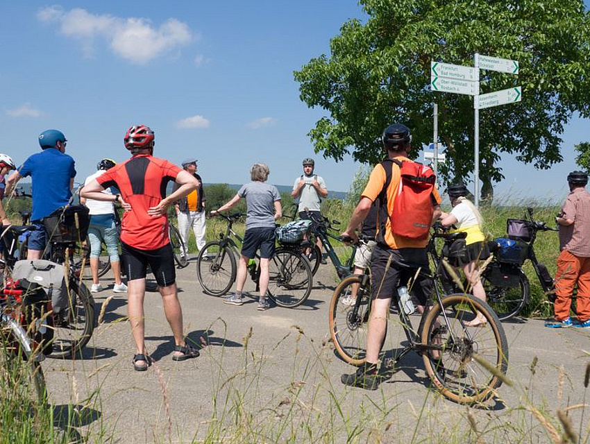 Eine Radfahrergruppe steht an einem Kreuzungspunkt zusammen und orientiert sich zur Weiterfahrt an der neuen Radwegebeschilderung.