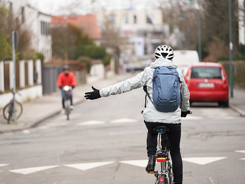 Blick auf eine städtische Straßensituation mit Radfahrern. Eine Person auf dem Rad im Vordergrund zeigt an, dass sie links abbiegen möchte.