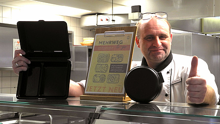 Ein Mann steht hinter dem Tresen einer Kantine. In der rechten Hand hält er eine Mehrwegverpackung, mit der linken Hand zeigt er mit dem Daumen nach oben.