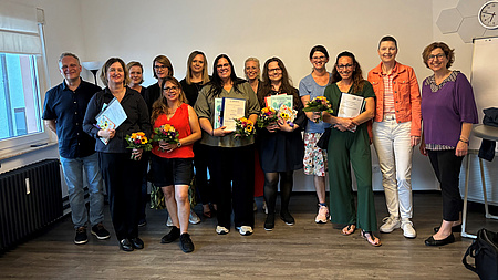 Zwölf Frauen und ein Mann stehen in einem Raum. Einige der Frauen halten Blumensträuße und Urkunden in Händen.