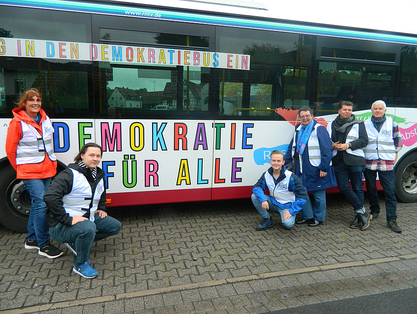 Bus mit Schriftzügen  „Demokratie für alle“ und „Steig in den Demokratiebus ein“. Davor einige Akteure