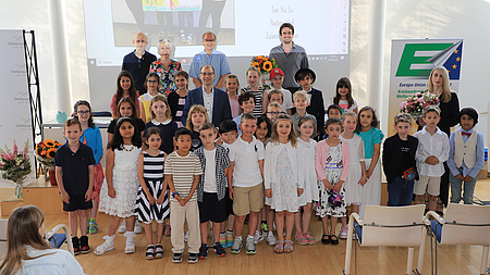 Zahlreiche Kinder und sechs Erwachsene stehen in einem Saal vor einer Leinwand.