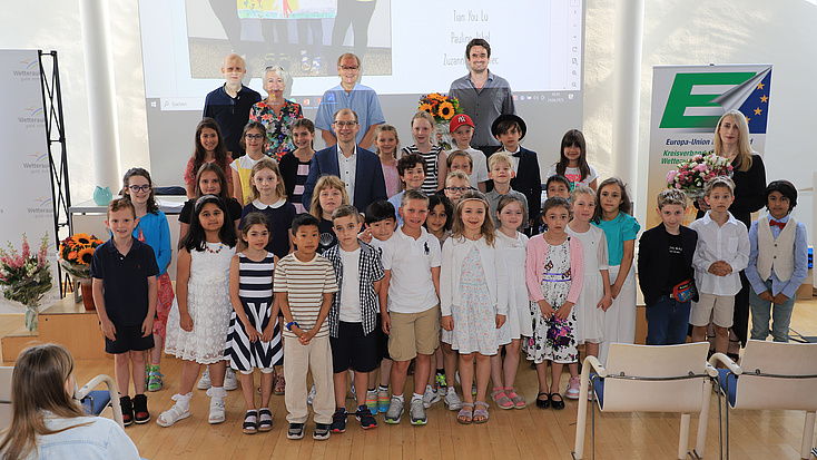 Zahlreiche Kinder und sechs Erwachsene stehen in einem Saal vor einer Leinwand.