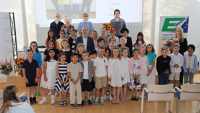Zahlreiche Kinder und sechs Erwachsene stehen in einem Saal vor einer Leinwand.