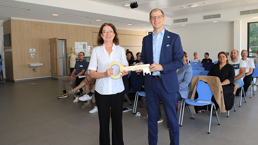 Eine Frau und ein Mann stehen in einem Saal und halten gemeinsam einen großen, goldenen Schlüssel in Händen. Im Hintergrund sitzen mehrere Menschen auf aufgestellten Stühlen.