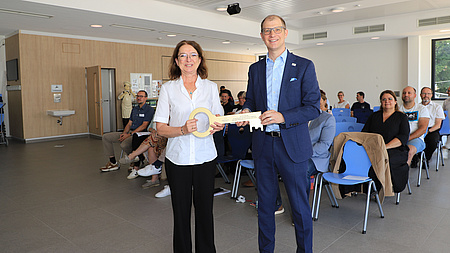 Eine Frau und ein Mann stehen in einem Saal und halten gemeinsam einen großen, goldenen Schlüssel in Händen. Im Hintergrund sitzen mehrere Menschen auf aufgestellten Stühlen.