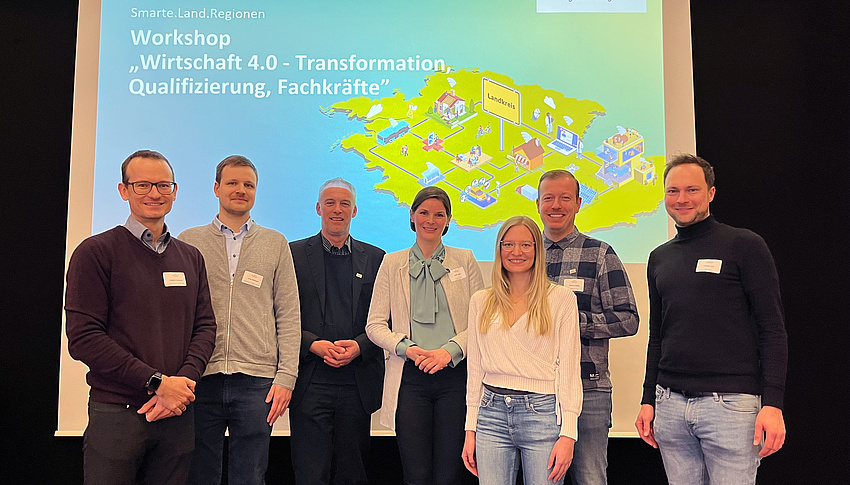 Zwei Frauen und fünf Männer stehen nebeneinander auf einer Bühne und lächeln in die Kamera. Im Hintergrund ein Ausschnitt aus der Präsentation des Workshops zu Wirtschaft 4.0