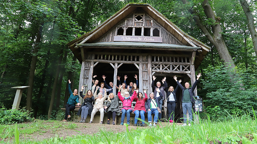 Zu sehen ist eine Hütte im Wald. Davor sitzt eine Gruppe von Leuten, die in die Kamera winkt.