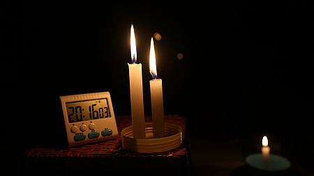 In einem dunklen Raum stehen auf einem Tisch drei brennende Kerzen neben einer Uhr mit digitaler Anzeige.