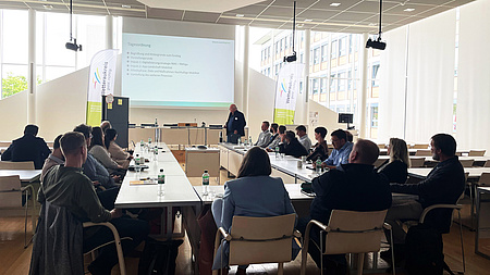 In einem Sitzungsraum sind Tische zu einem Rechteck angeordnet. Daran sitzen mehrere Personen.- Im Vordergrund eine Leinwand, auf der eine Präsentation des Rhein-Main-Verkehrsverbundes zu sehen ist.