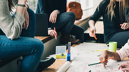 4 Frauen, die um einen niedrigen Tisch sitzen. Sie arbeiten zusammen und machen Notizen auf Papieren, die auf dem Tisch liegen.