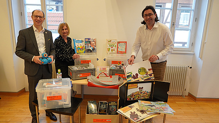 Eine Frau und zwei Männer stehen in einem größeren Raum vor einem Tisch, auf und vor dem verschiedene Bücher und weitere Medien platziert sind. Alle drei halten zudem Bücher und weitere Medien in den Händen.