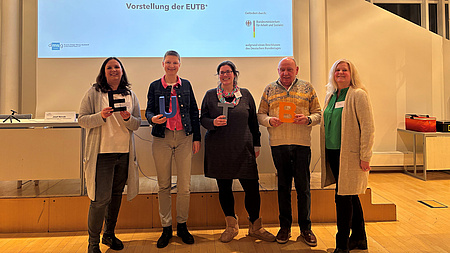 Vier Frauen und ein Mann stehen in einem großen Saal und halten die Buchstaben E, U, T und B in Händen.