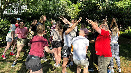 Eine größere Gruppe Menschen führt auf einer Wiese im Freien eine gemeinsame, koordinierte Aktivität aus.