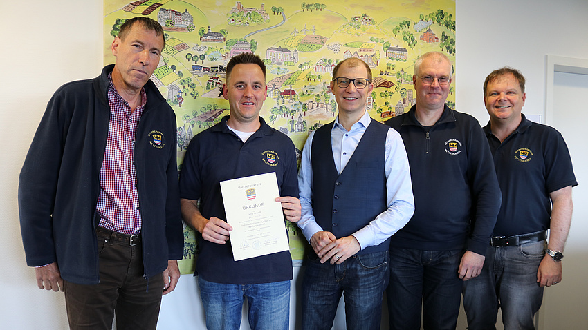 Fünf Männer stehen nebeneinander und lächeln in die Kamera. Einer der Männer hält ein Schriftsück in der Hand.