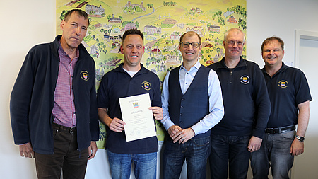 Fünf Männer stehen nebeneinander und lächeln in die Kamera. Einer der Männer hält ein Schriftsück in der Hand.