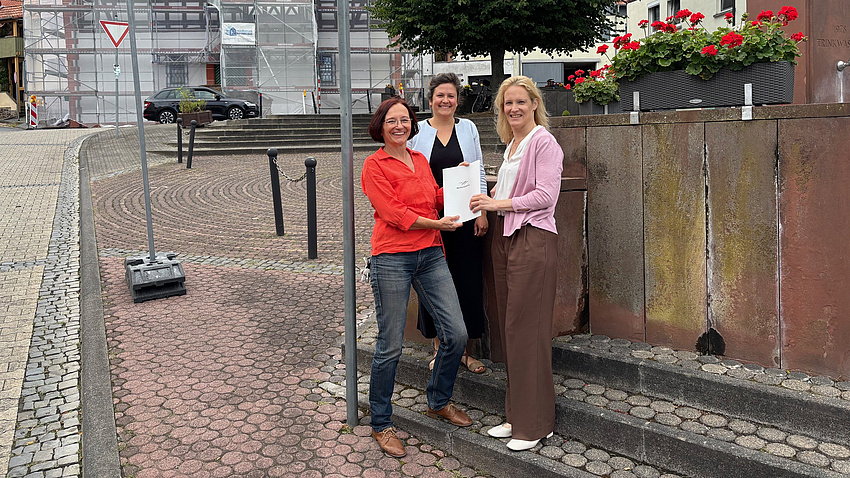 Drei Frauen stehen auf einem Platz vor einer Sandsteinmauer. Zwei der Frauen halten ein Dokument in Händen.