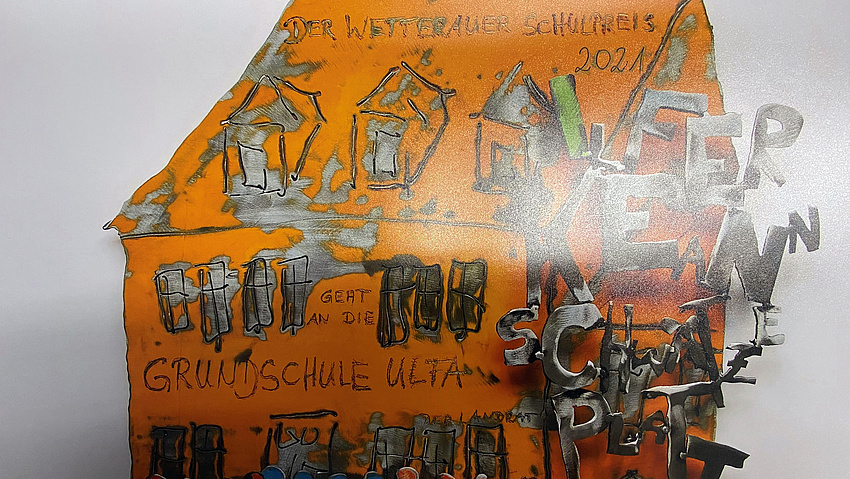 Eine Metallskulptur, die ein Schulgebäude darstellt. Darauf steht: Der Wetterauer Schulpreis 2021 geht an die Grundschule Ulfa..