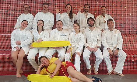 Elf Männer und Frauen sitzen in zwei Reihen hintereinander. Vor ihnen auf dem Boden liegt eine Frau auf der rechten Seite.