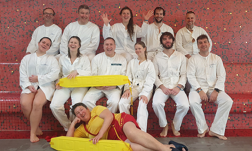 Elf Männer und Frauen sitzen in zwei Reihen hintereinander. Vor ihnen auf dem Boden liegt eine Frau auf der rechten Seite.