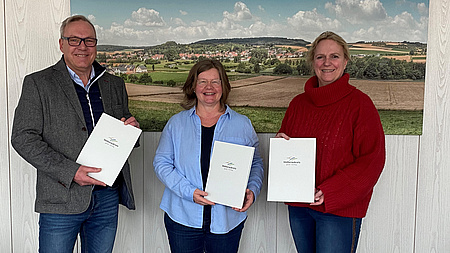 Die drei Personen halten eine Urkunde in der Hand.